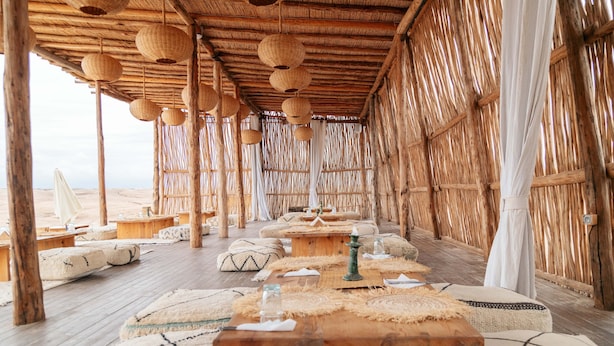 A thatched-roof room with wooden furniture, hanging lanterns, and a view of the beach. - Alkamar Camp Agafay (Agafay)