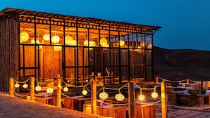 A wooden structure with large windows and string lights, surrounded by tables and chairs. - Alkamar Camp Agafay (Agafay)