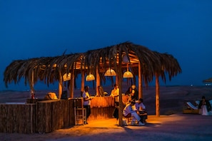 A thatched-roof outdoor seating area with a bar, surrounded by palm trees, under a night sky. - Alkamar Camp Agafay (Agafay)