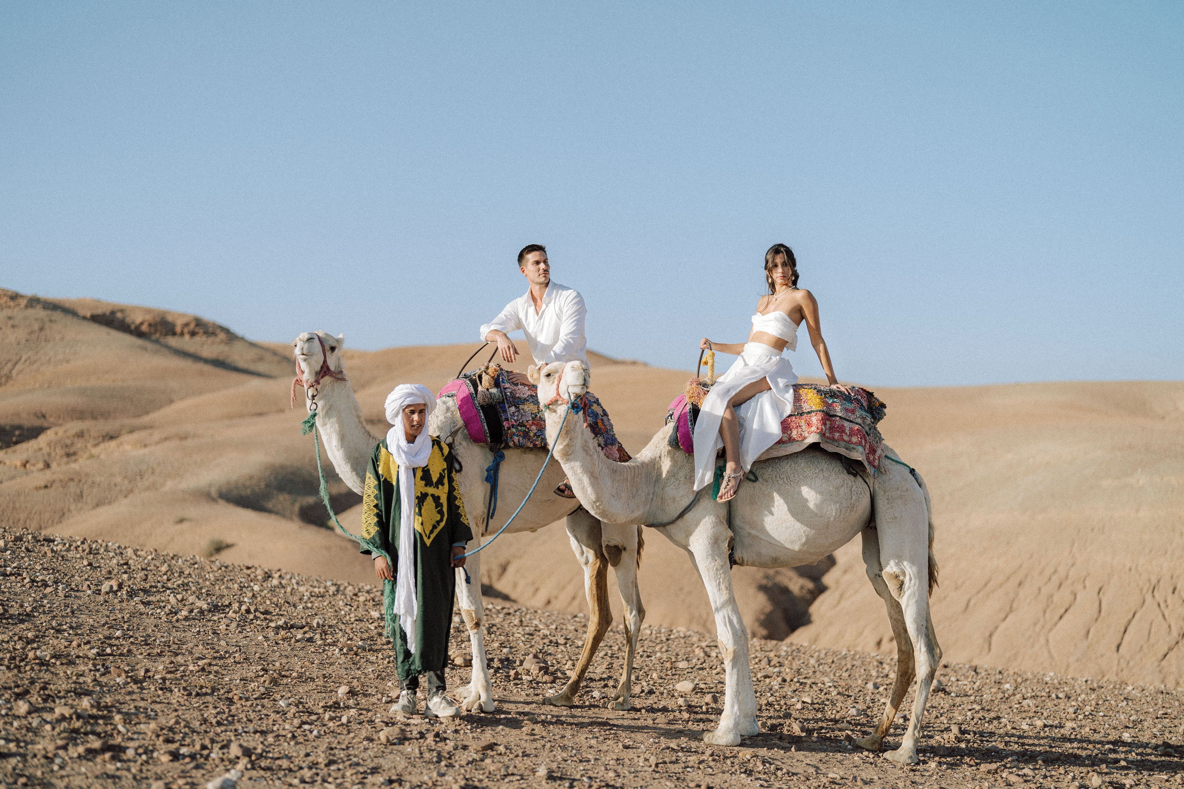 Paisaje desértico con tres camellos, uno llevando a una persona vestida de blanco y otro con una persona en un vestido blanco.