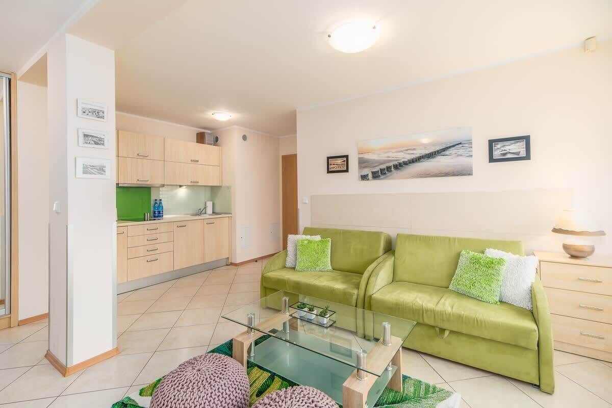 Un salon moderne avec un canapé vert, une table basse en verre et une cuisine en arrière-plan.