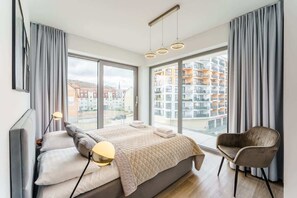 Un dormitorio moderno con una cama grande, una silla y vistas a un paisaje urbano a través de amplios ventanales.
