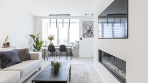 Un salon moderne avec un canapé, une table basse et des fauteuils. On y trouve une cheminée, un coin repas avec une table et des chaises, ainsi qu’une grande fenêtre avec des stores.