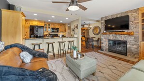 A spacious living area with a fireplace, a ceiling fan, and a kitchen in the background.