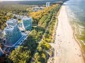 Vue aérienne d’une zone côtière avec des bâtiments modernes, une plage de sable et une forêt dense.