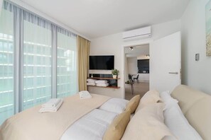 Une chambre d’hôtel avec un grand lit, une télévision, une petite table avec une plante et une vue sur la ville grâce à de grandes fenêtres.