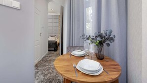 En matplats med ett runt träbord dukat för två personer, en vas med blommor och en utsikt över ett sovrum i bakgrunden.