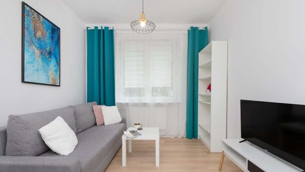 A modern living room with a grey sofa, a white coffee table, a flat-screen TV on a white stand, teal curtains, and a large abstract painting on the wall. - Comfortable Apartment with Bedroom | Work Space (Gdańsk)