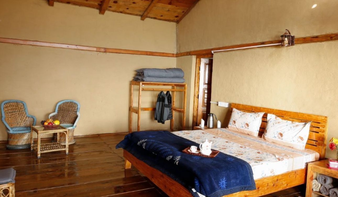 A wooden bed with a blue blanket, two wicker chairs, a small table with fruit, and a shelf with folded clothes.