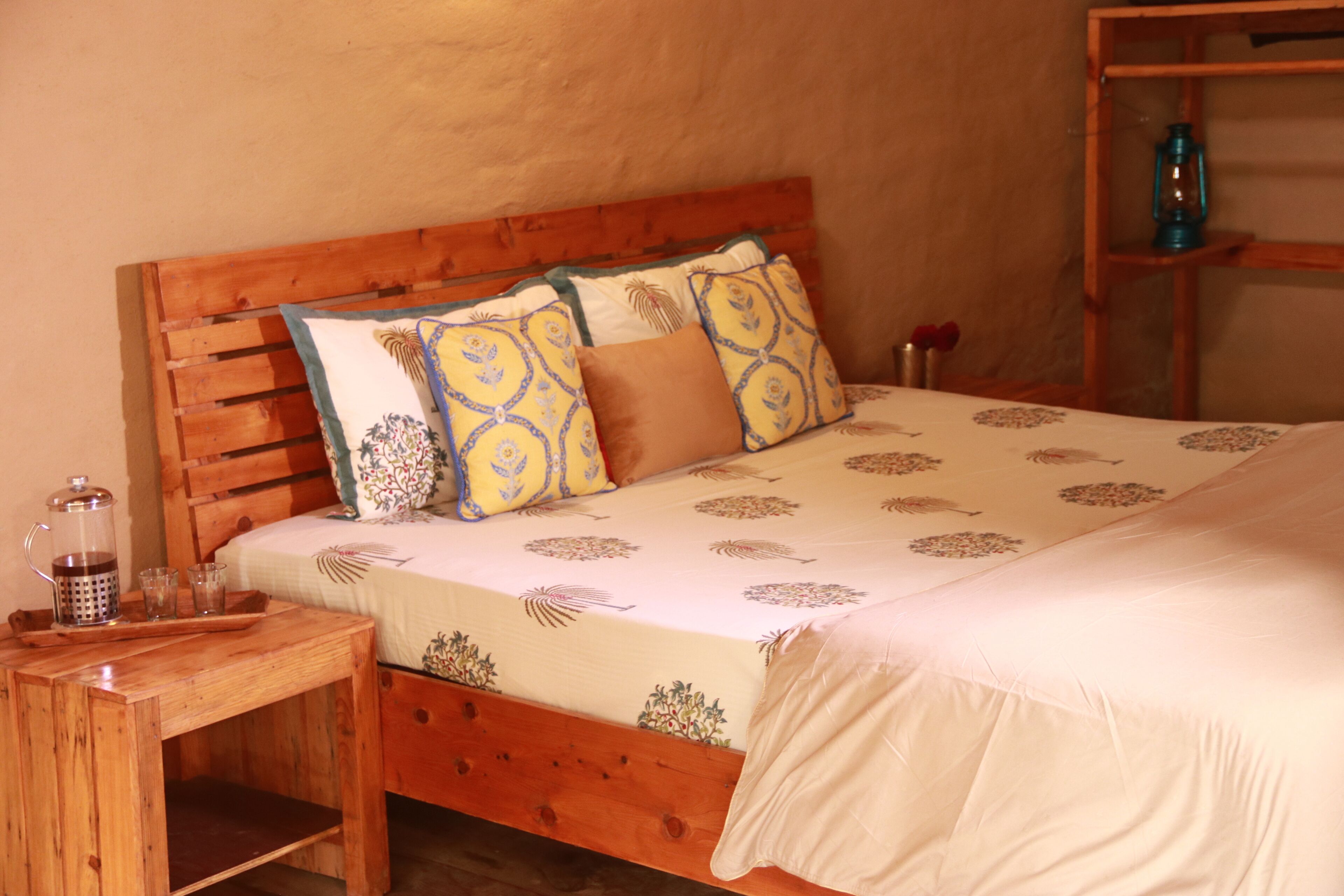 A wooden bed with a patterned bedspread and pillows, a wooden nightstand with a kettle, and a lantern on a shelf.
