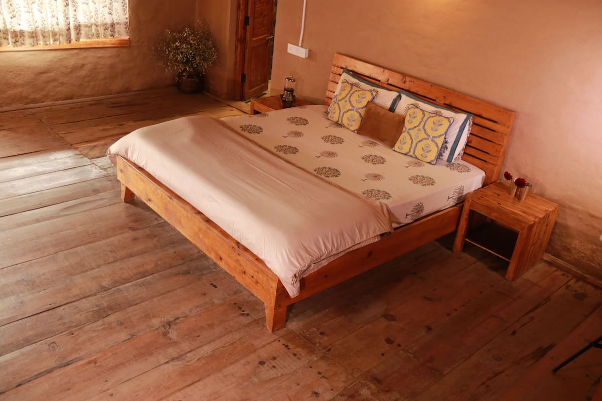 A wooden bed with a beige bedspread and patterned pillows, a small wooden nightstand with a vase of flowers, and a window with curtains.
