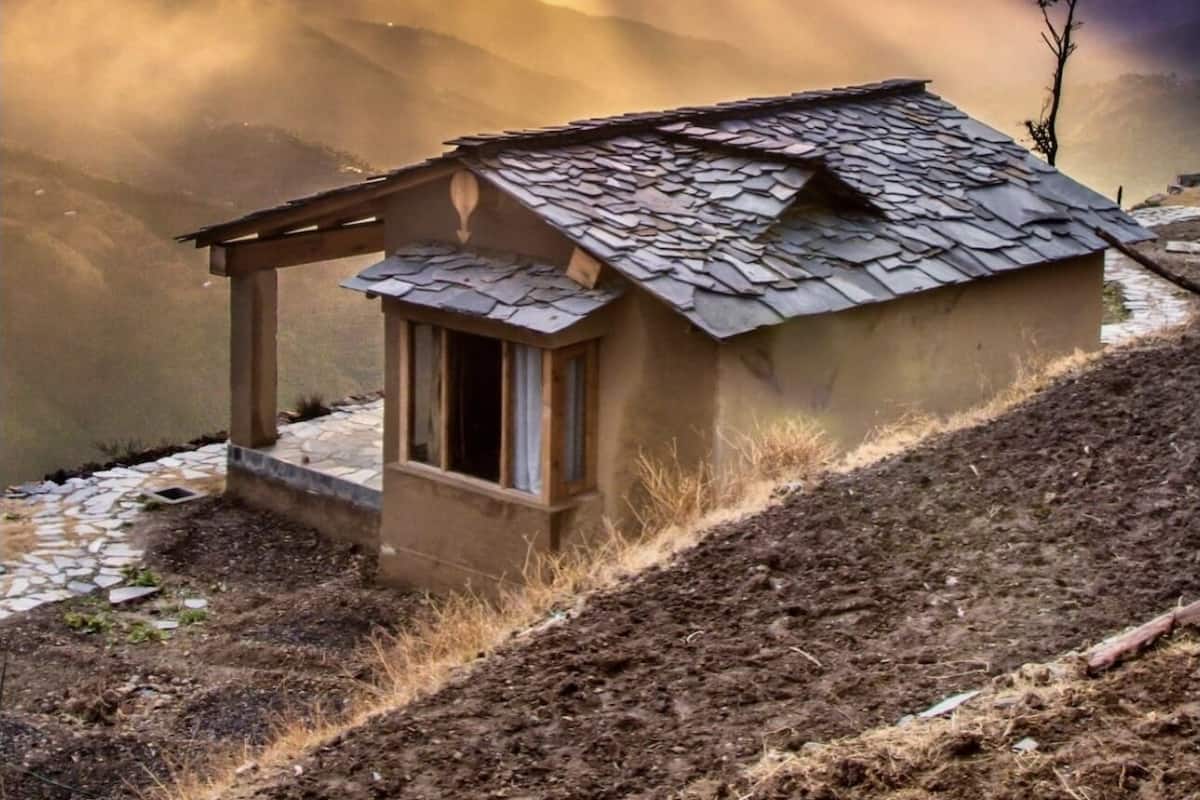 A small, rustic cabin with a sloped roof, situated on a hillside with a view of distant mountains and a dramatic sky.