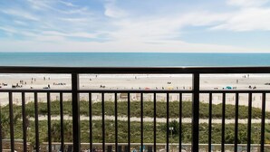 A beach view from a balcony with a railing, beach in the background, and some greenery in the foreground.