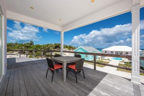 A covered outdoor area with a wooden deck, a table with chairs, and a view of a beach and buildings.