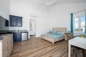 A bedroom with a wooden floor, a bed with a blue patterned bedspread, a bedside table with a lamp, and a window with a view.