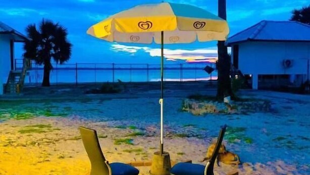 Escena en la playa con una mesa y sillas bajo un paraguas grande, palmeras y un edificio al fondo.