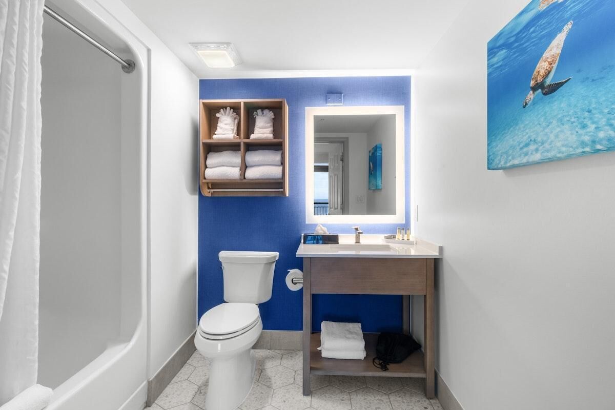 Een badkamer met een wit toilet, een houten plankje met handdoeken, een wastafel met spiegel en een schilderij van een zeeschildpad.