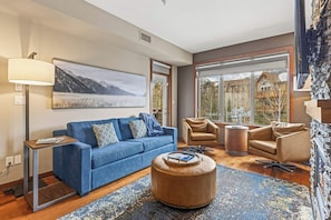 A modern living room with a blue sofa, two armchairs, a round ottoman, and a large window offering a view of a mountain and a house.