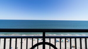 Vista da praia a partir de uma varanda com corrimão, céu claro e oceano.
