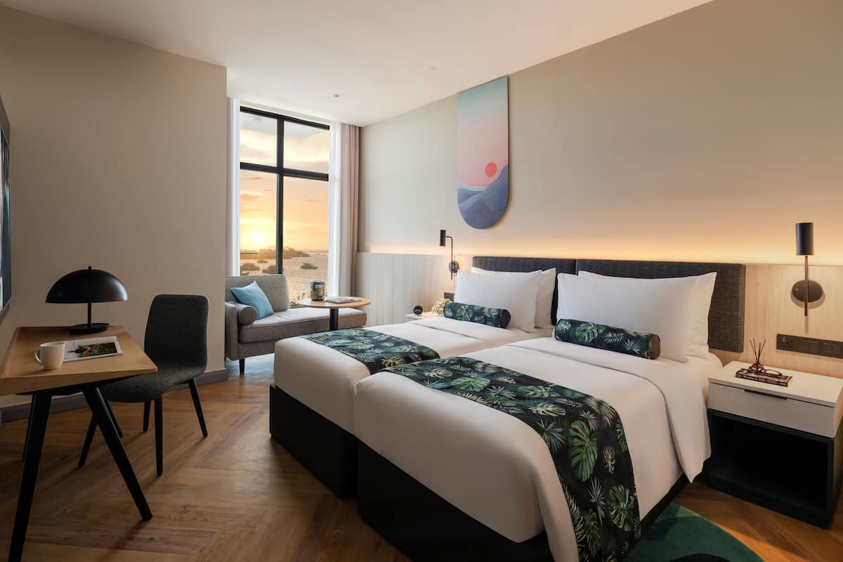 A hotel room with a large bed, a desk, a chair, and a view of the sunset through the window.