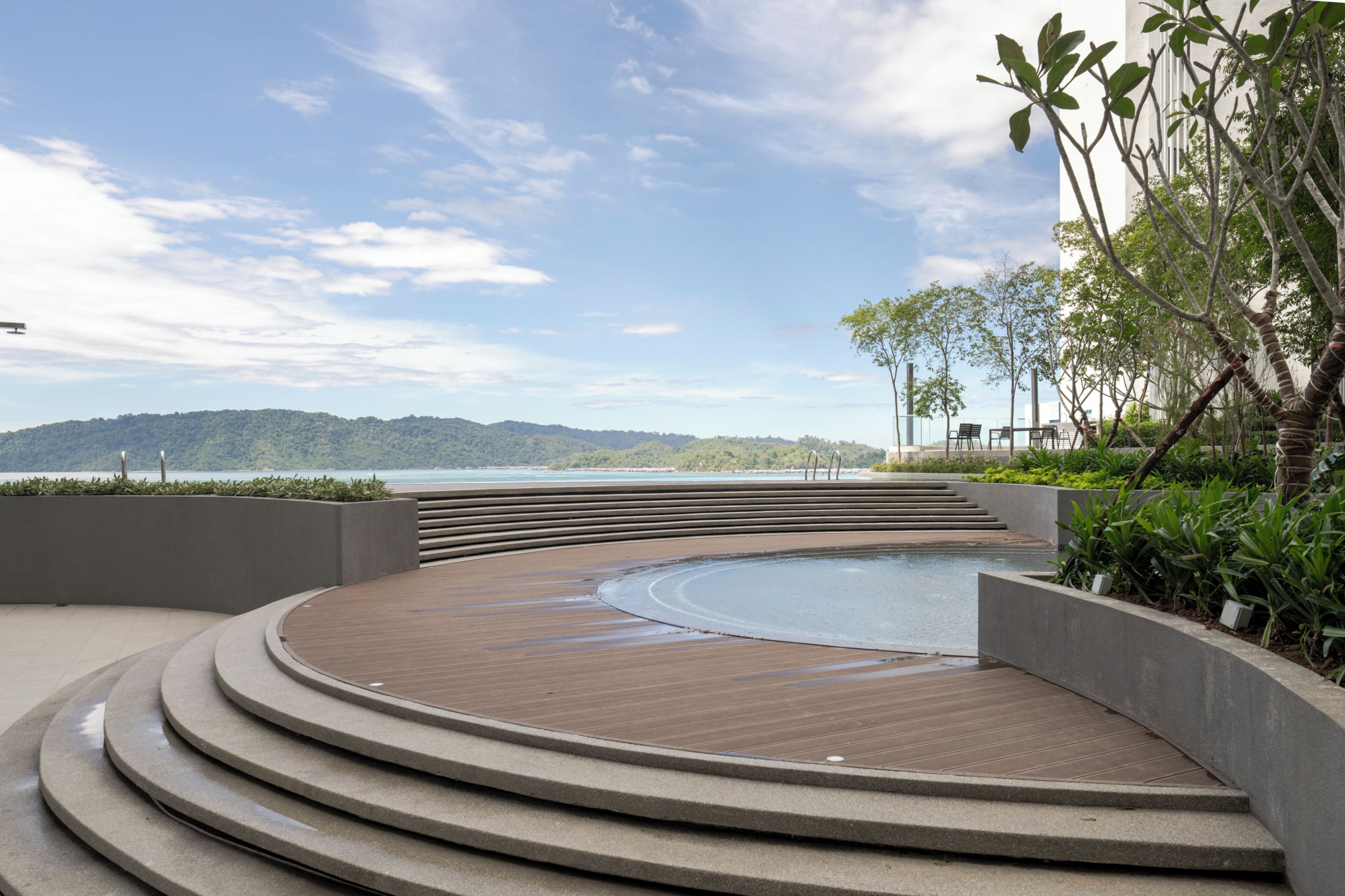 Une terrasse circulaire en bois, dotée d’une piscine, entourée d’escaliers en béton et de verdure, et offrant une vue sur une étendue d’eau et des collines lointaines.
