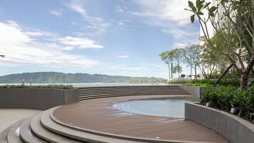 A circular wooden deck with a pool, surrounded by concrete steps and greenery, overlooking a body of water and distant hills.