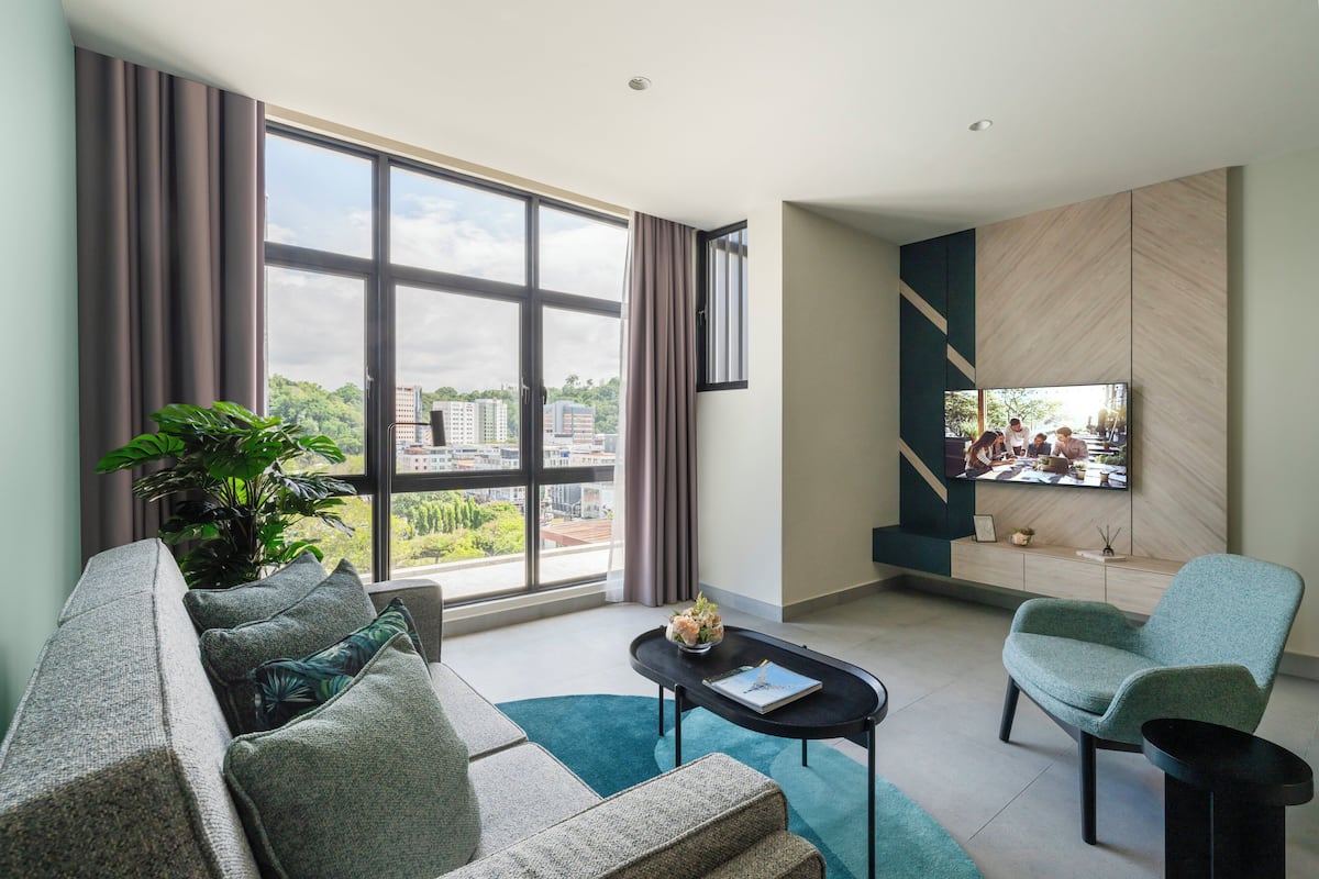 A modern living room with a sofa, armchair, and a coffee table. A large window offers a city view.