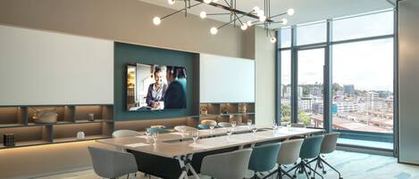 A modern conference room with a long table, chairs, a large screen, and a city view through large windows.