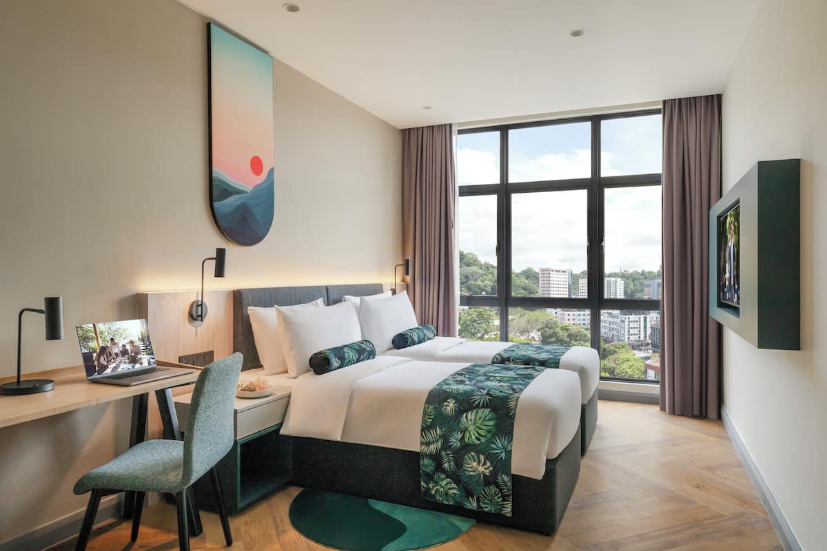 A modern hotel room with a large bed, a desk with a laptop, a chair, and a view of the city through large windows.