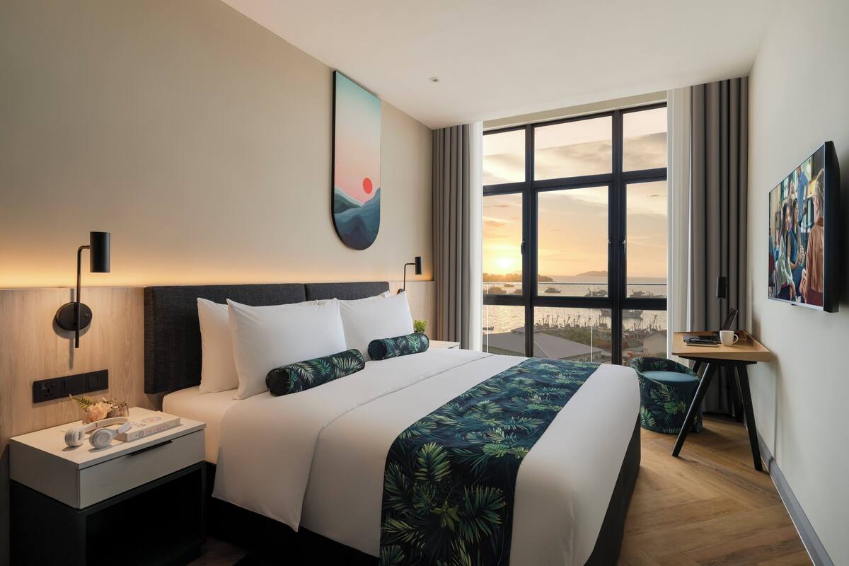 A modern hotel room with a large bed, a nightstand with a lamp, a desk with a chair, and a large window offering a city view.