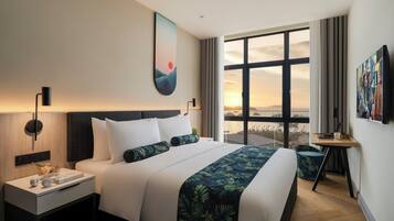 A modern hotel room with a large bed, a nightstand with a lamp, a desk with a chair, and a large window offering a city view.