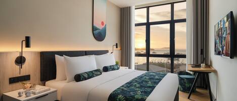 A modern hotel room with a large bed, a nightstand with a lamp, a desk with a chair, and a large window offering a city view.