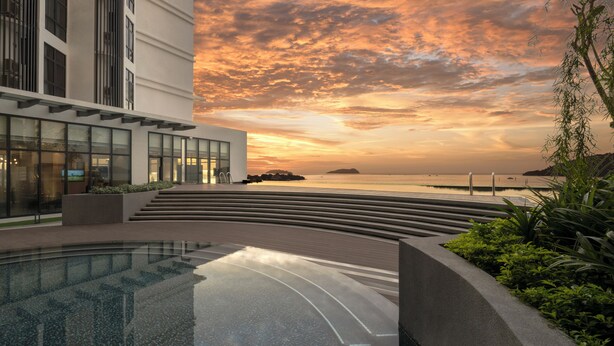 A modern building with a glass facade, a pool area with a curved edge, and a view of the sunset over the water. - Citadines Waterfront Kota Kinabalu (Kota Kinabalu)