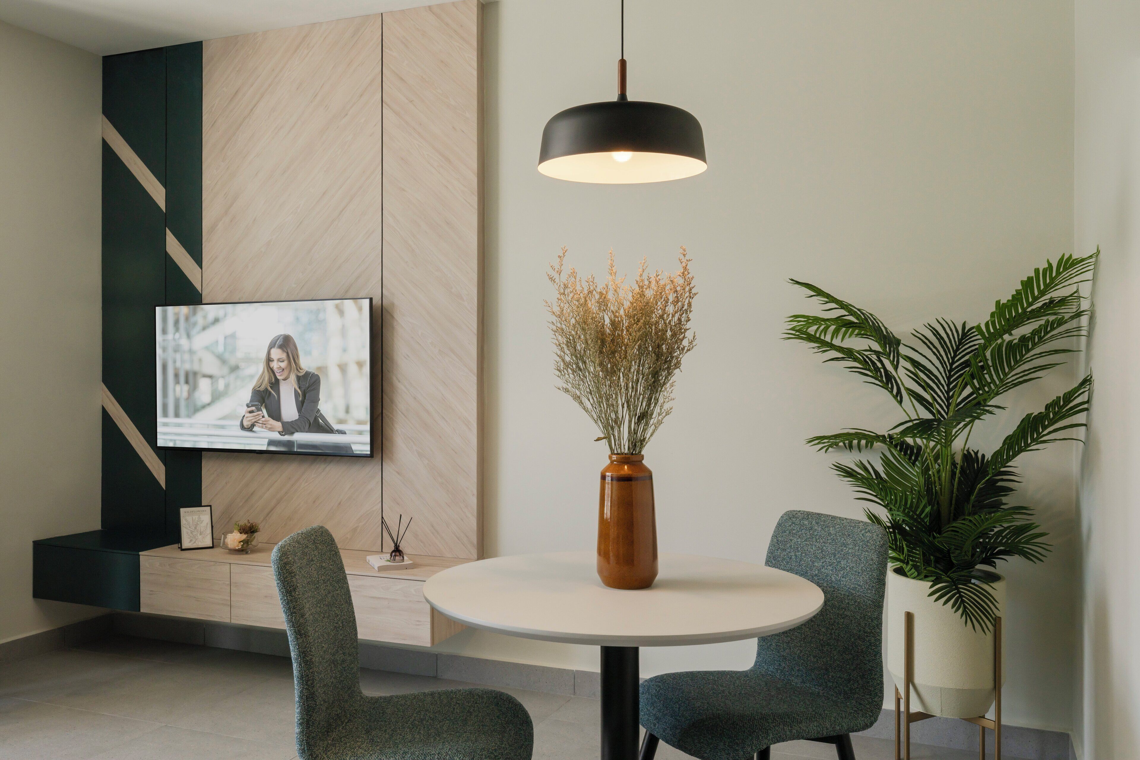 Un coin repas moderne avec une table ronde, deux chaises, un vase contenant des fleurs séchées, une grande plante en pot et un téléviseur fixé au mur.