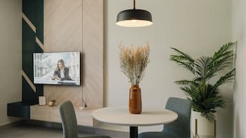 A modern dining area with a round table, two chairs, a vase with dried flowers, a large potted plant, and a television mounted on the wall.