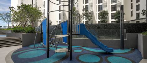 An outdoor playground with a slide and climbing structure, surrounded by a patterned carpet and a stone wall.