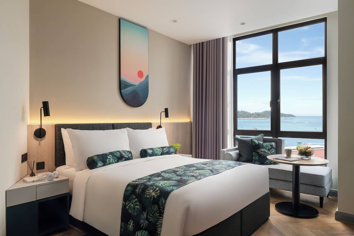 A modern hotel room with a large bed, a side table, a small round table, and a view of the sea through large windows.