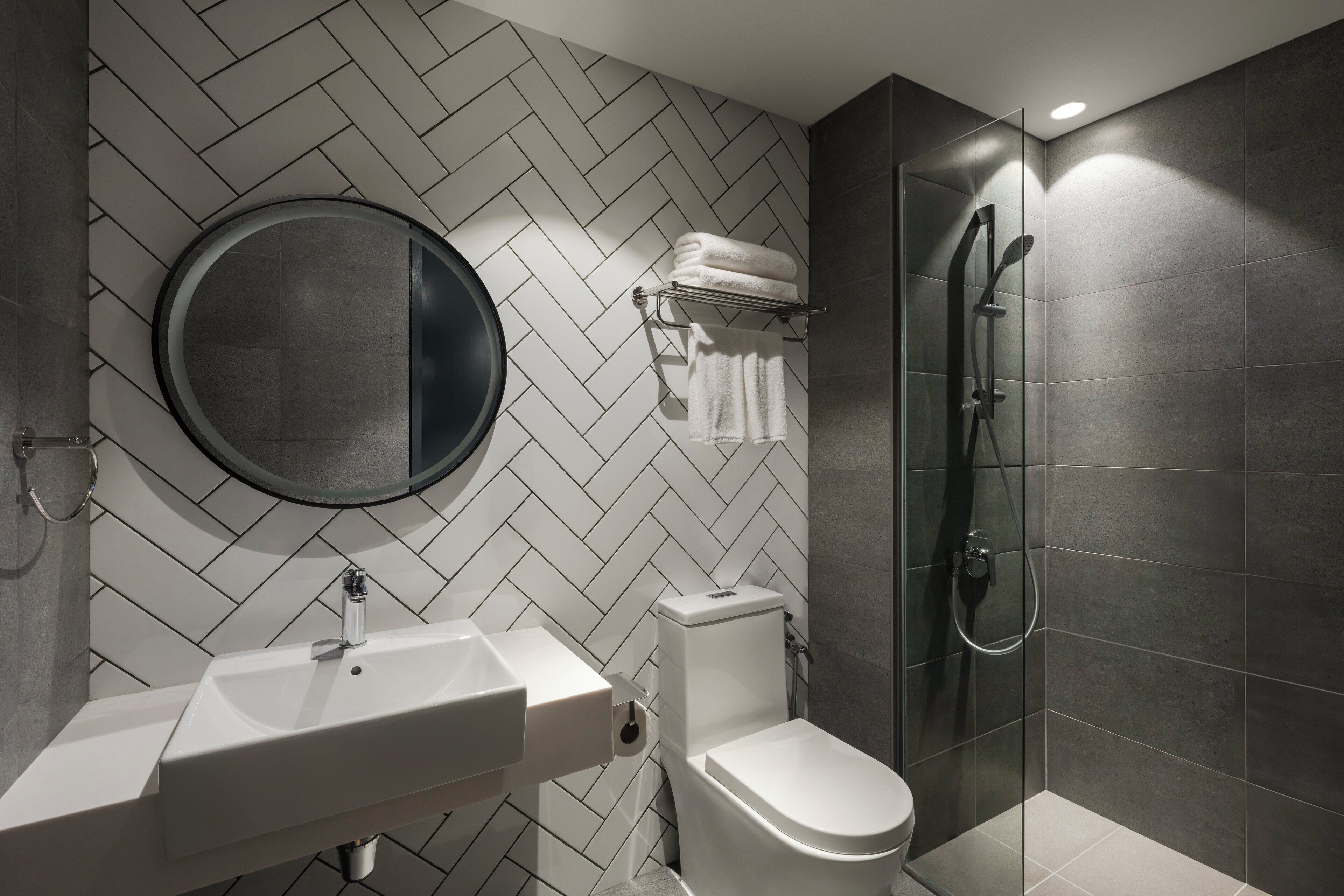 Een moderne badkamer met een backsplash van visgraat tegels, een ronde spiegel, een witte wastafel, een toilet en een douchecabine met glazen wanden.