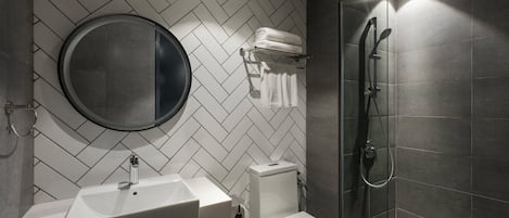 Un baño moderno con un resalto de azulejos en patrón espiga, un espejo redondo, un lavamanos blanco, un inodoro y una ducha con mampara de vidrio.