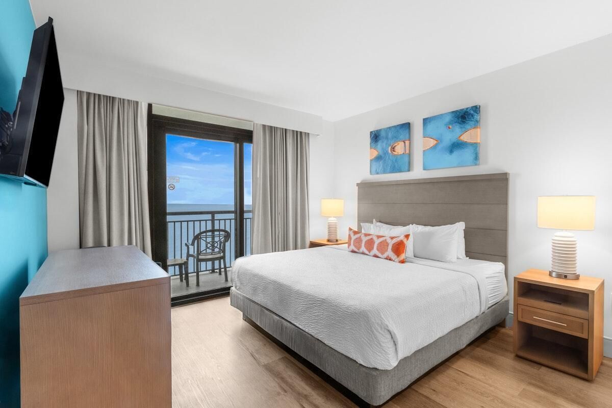 Habitación de hotel con cama, dos lámparas de noche, televisor montado en la pared, escritorio y balcón con una silla.