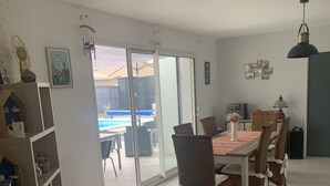 Et spiseområde med et bord, stole, en reol og udsigt til en swimmingpool gennem glasdøre.