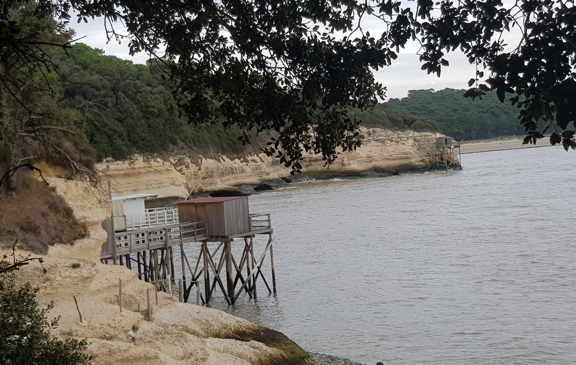 Un quai au bord de la rivière, avec une petite construction en bois, entouré d’arbres et bordé d’une plage de sable.