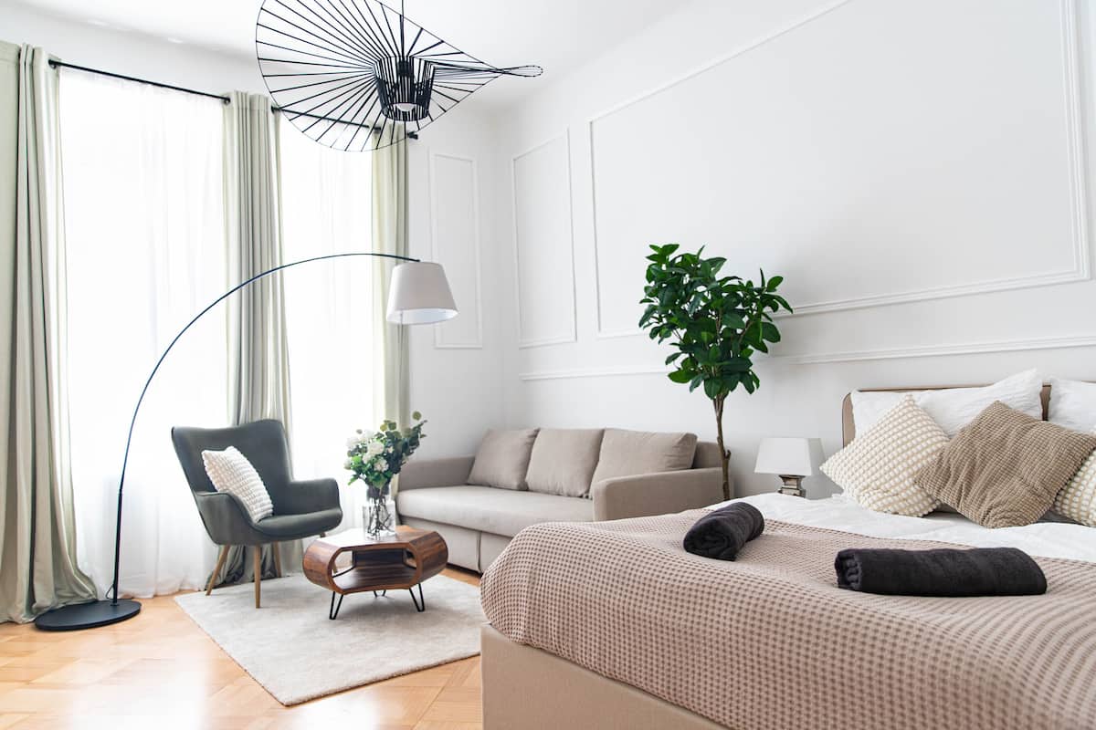 Une chambre d’hôtel moderne comprenant un lit, un canapé, une chaise, une lampe sur pied, une table avec un vase et une plante en pot.