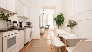A modern kitchen with white cabinetry, a built-in oven, and a washing machine. There is a dining area with white chairs and a table, and a plant on the windowsill. - City Apartment - 5 min to Metro (Vienna)