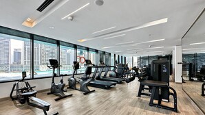 A modern gym with various exercise equipment, large windows with a city view, and a clean, spacious layout. - WelHome - Contemporary 1BR in Binghatti Canal & Burj Views (Dubai)