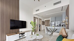 A modern living room with a flat-screen TV, a white entertainment unit, a dining area with a glass table, and a kitchen in the background. - WelHome - Contemporary 1BR in Binghatti Canal & Burj Views (Dubai)
