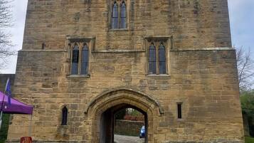A stone castle entrance with a central archway, a flag, and a sign indicating a tourist attraction.