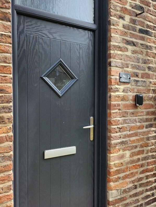 A dark grey door with a diamond-shaped window and a brass handle, set in a brick wall with a sign reading "No. 2 The Snug". - 1 Bedroom - Sleeps 4 - Pets - Parking on Site (Morpeth)