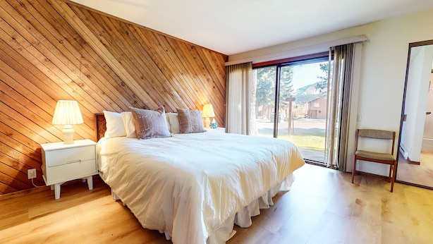 A bedroom with a large bed, wooden panel walls, a nightstand with a lamp, and a view through a window.