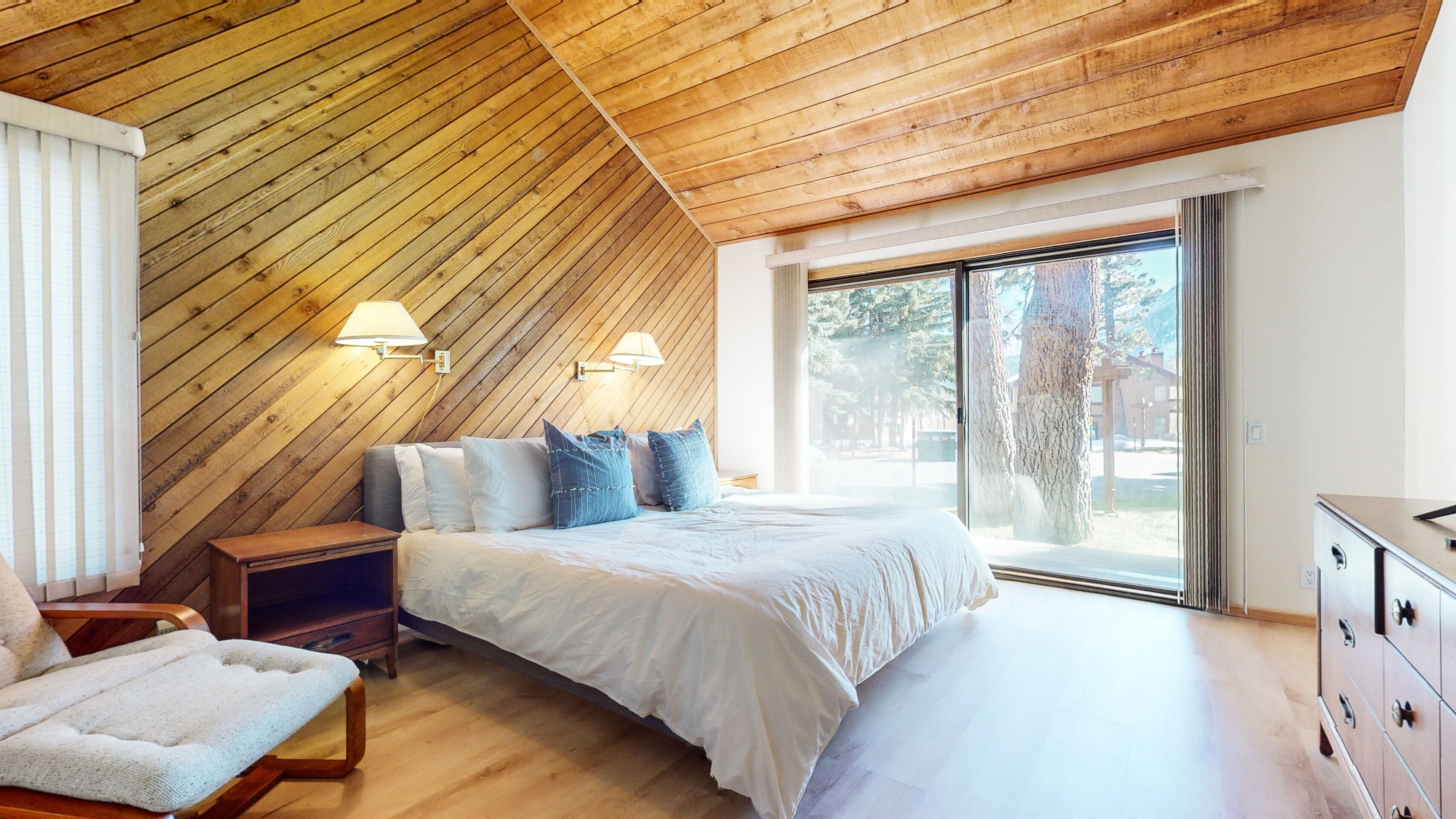 A bedroom with a sloped wooden ceiling, a bed with white bedding, two bedside lamps, a wooden nightstand, a chair, and a dresser.